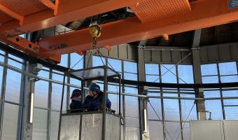 Zwei Fachkräfte mit Schutzhelmen stehen in einer Arbeitsbühne in dem technischen Bauwerk. Um sie herum verläuft ein Gerüst, daneben ist ein großes orangefarbenes Hebezeug mit Kranhaken zu sehen. Die Umgebung wirkt industriell.