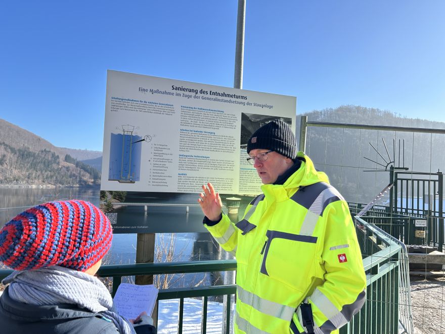 Zwei Personen stehen vor einer großen Informationstafel zur Sanierung des Entnahmeturms. Eine Person in leuchtend gelber Warnschutzjacke erklärt Inhalte der Tafel, während die andere Person Notizen macht. Im Hintergrund ist die Talsperre und das Wasser