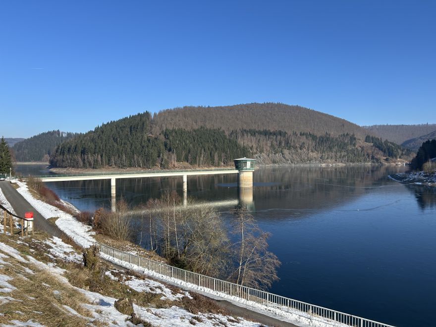 Panorama der Talsperre Schönbrunn im Winter: Ein ruhig spiegelnder Stausee, durchzogen von einer schmalen Brücke, an deren Ende der Entnahmeturm steht. Im Hintergrund bewaldete Hügel, im Vordergrund verschneite Uferbereiche und ein Gehweg.