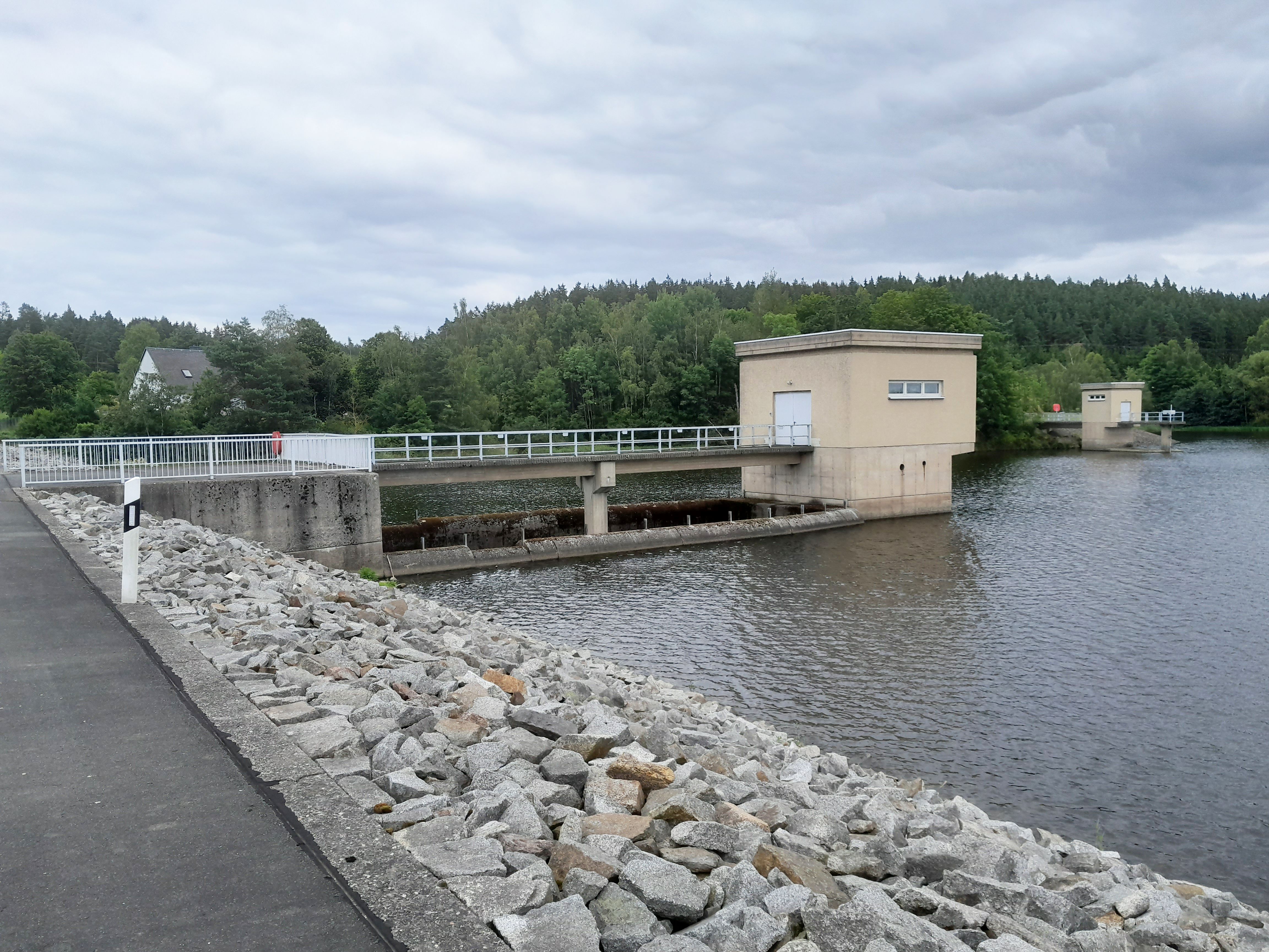 Im Vordergrund liegt der mit Schüttsteinen befestigte Damm, dahinter ragt das Abschlussbauwerk mit der Hochwasserentlastung in die Wasserfläche.