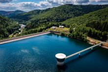 Luftaufnahme mit Blick auf den gefüllten Stauraum, der Wasserentnahmeturm im Stauraum ist umgeben mit blauen Wasser, im Hintergrund kann man die Ortschaft sehen und das Dienstgebäude