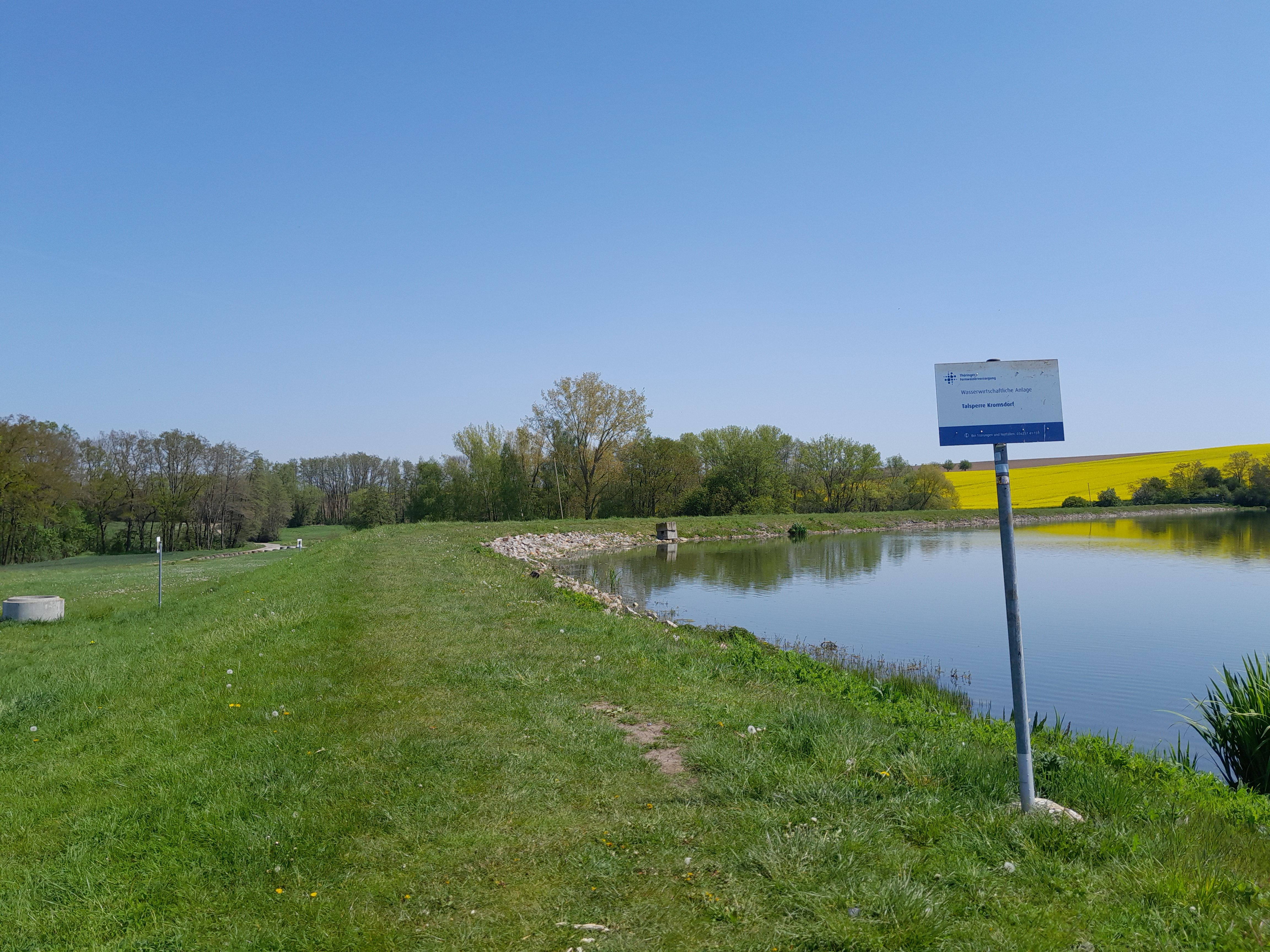 grüner Damm links und rechts die Wasserfläche der Stauanlage. Im Vordergrund ist das Schild mit dem Namen der Talsperre Kromsdorf und dem Betreiber TFW.
