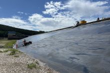 Bauarbeitende mit einer Maschiene bringen auf der wasserseitigen Dammschicht den Asphalt auf. Das Becken ist entleert.