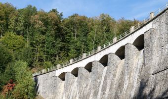 Ansicht einer gemauerten Talsperre mit bogenförmigen Stützpfeilern. Die Staumauer erstreckt sich entlang eines bewaldeten Hangs. Über der Anlage ist ein blauer Himmel mit dünnen Wolken zu sehen.