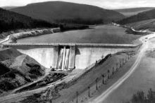 zu sehen ist in schwarz/weiß die neu errichtete Staumauser mit gefülltem Stauraum, herum ist die Infrastruktur noch am Entstehen