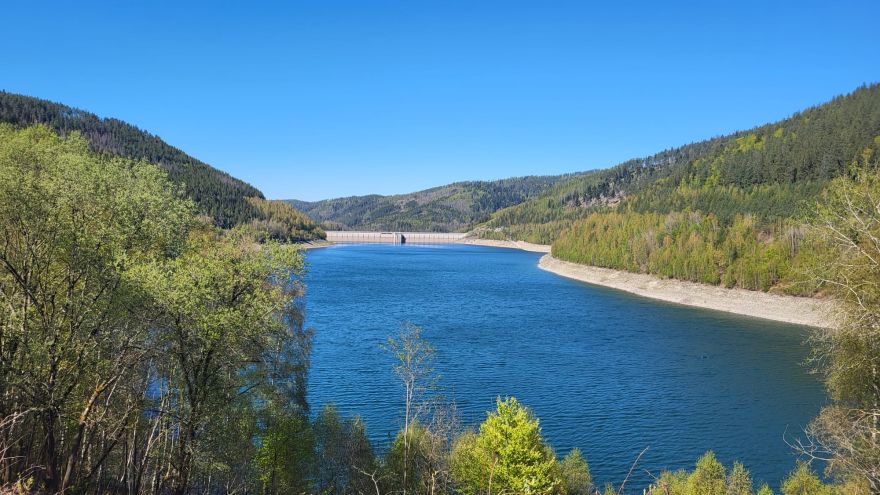 im Hintergrund ist die Staumauer, im Vordergrund ist der gefüllte Stauraum mit blauem Wasser und links und rechts sind die bewaldeten Hänge des Stauraums