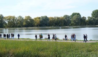 Mehrere Personen gehen auf einem Weg entlang eines Hochwasserrückhaltebeckens. Im Hintergrund sind die Wasserfläche des Beckens sowie ein Baumgürtel zu sehen. Im Vordergrund befindet sich eine Wiese.