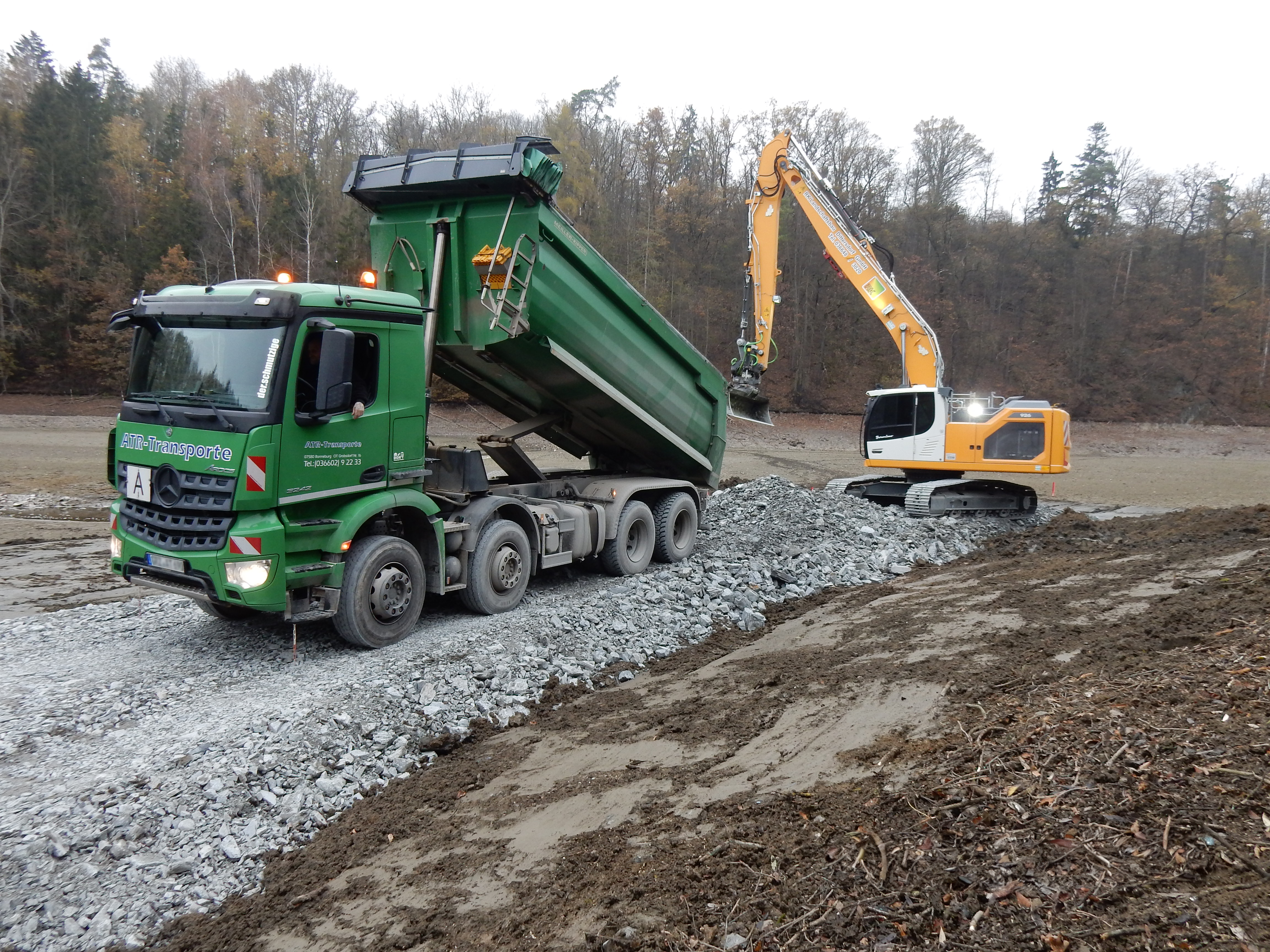 Ein Lastwagen bringt die Schüttung für die Errichtung der Baustraße und wird von einem Bagger entladen.