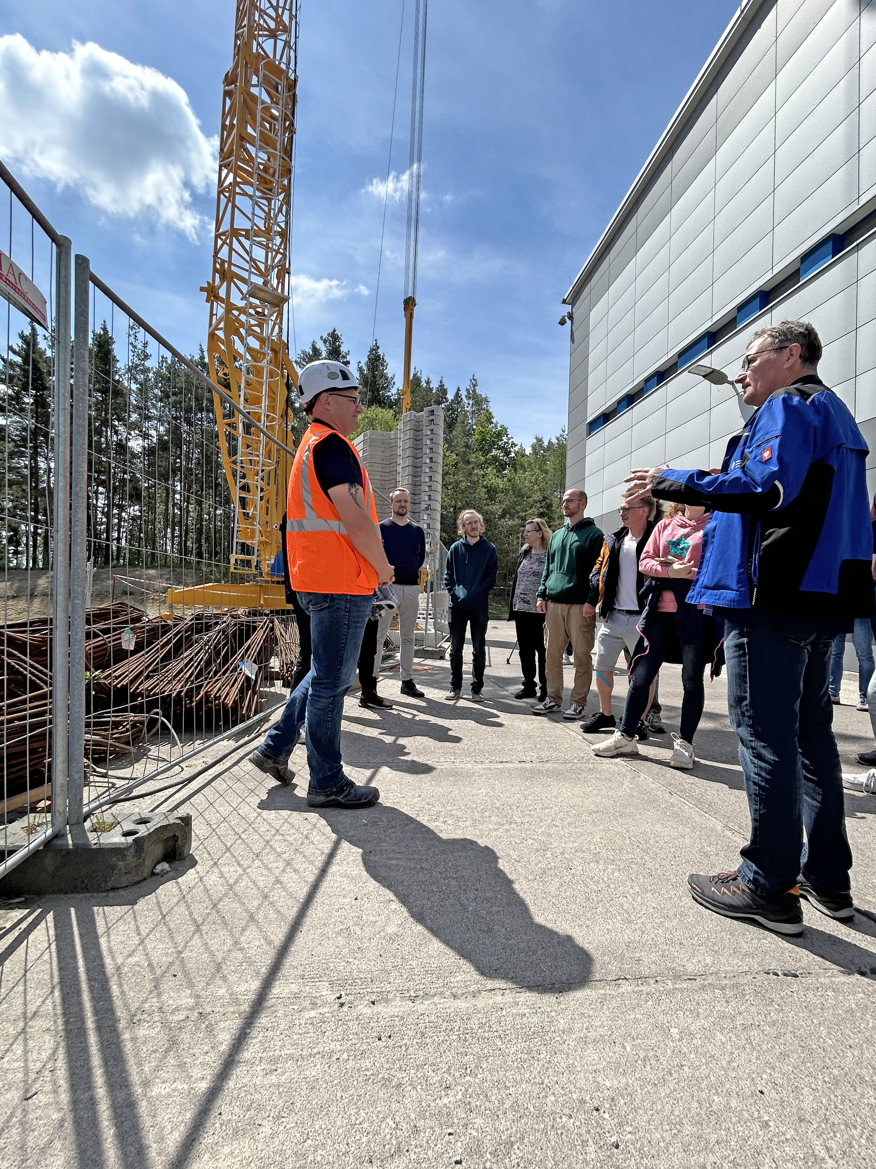 Bauleiter mit Warnweste in der Gesprächssitzuation mit mehreren Studierenden vor der Baustelle. Im Hintergrund sind der Kran und die Bewährungsstangen zu sehen.