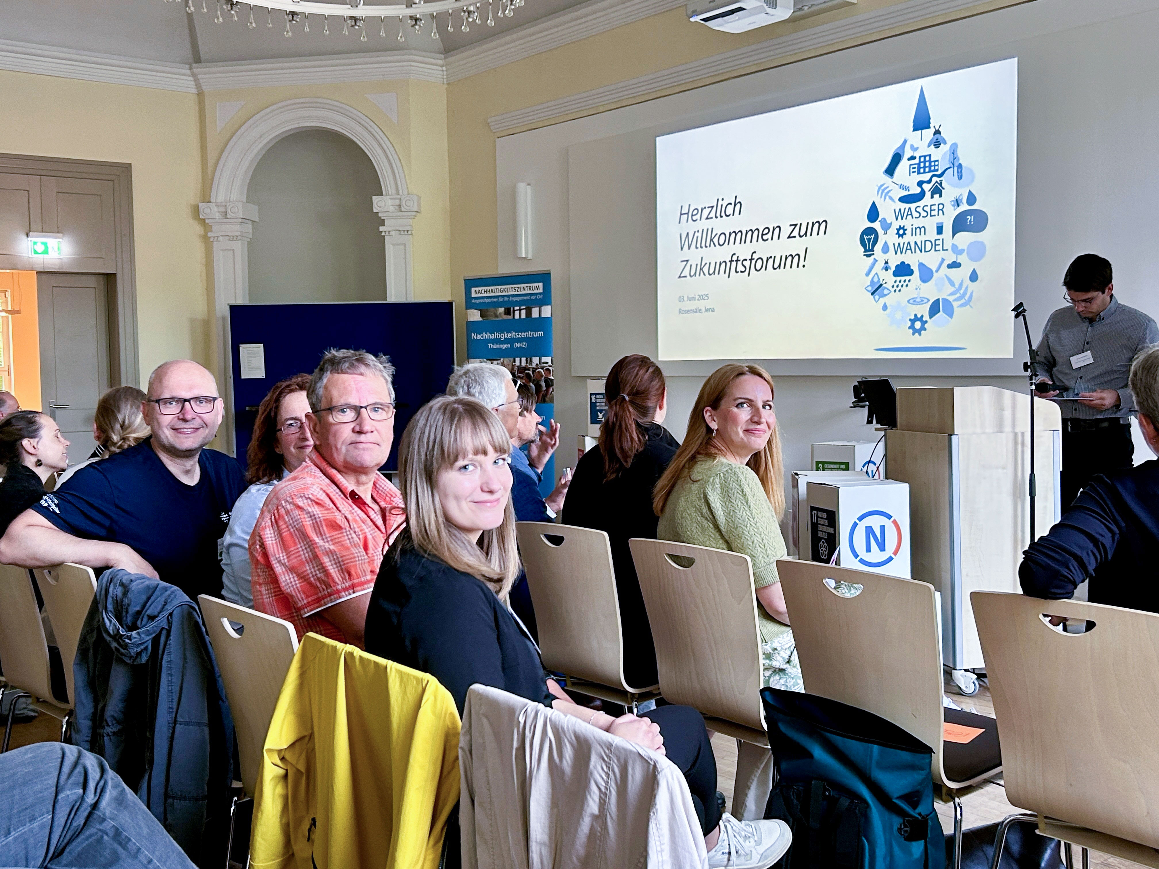Kollegen und Kolleginnen der TFW sitzen in einem Saal und blicken über die Schulter in die Kamera, dahinter ist auf einer Leinwand der Titel der Veranstaltung zu sehen.