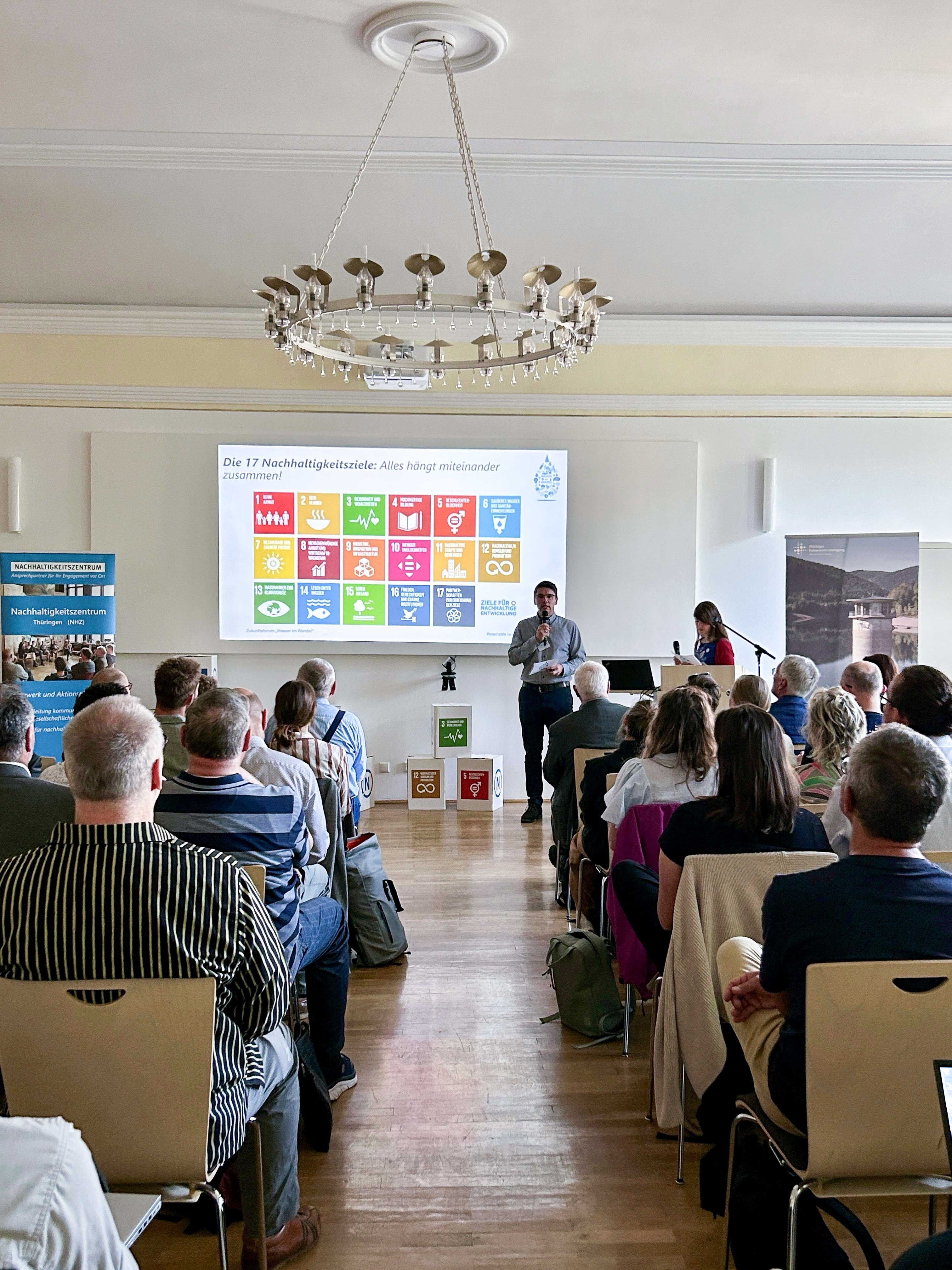 Totale eines gefüllten Veranstaltungssaals mit Redner und Leinwand, auf der die globalen Nachhaltigkeitsziele dargestellt sind.