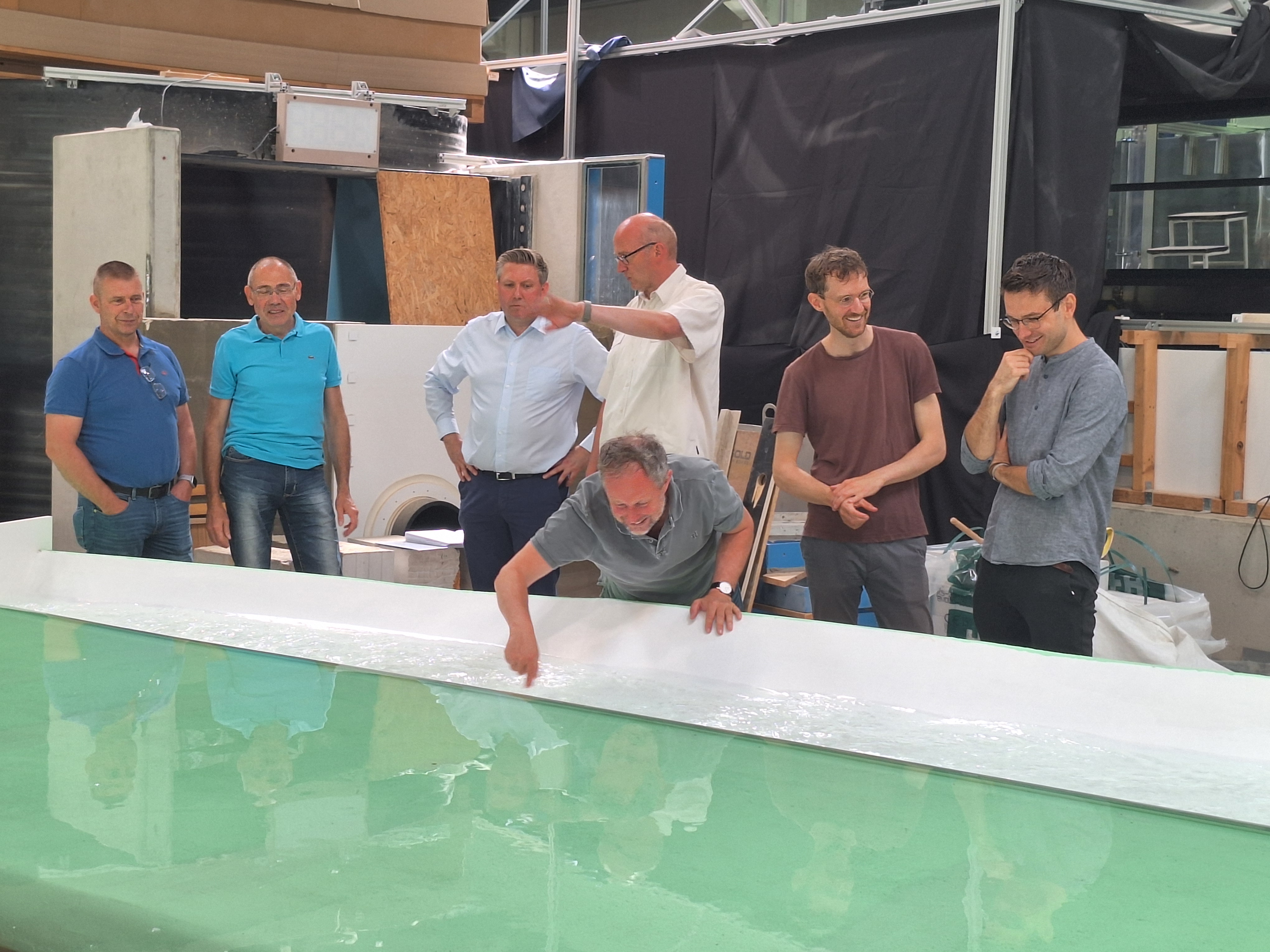 Menschen unterhalten sich im Hintergrund eines mit Wasser gefüllten Miniaturmodells des Hochwasserrückhaltebeckens Straußfurt, während ein Vertreter des beauftragten Planungsbüros die Wassertemperatur mit dem Finger prüft.