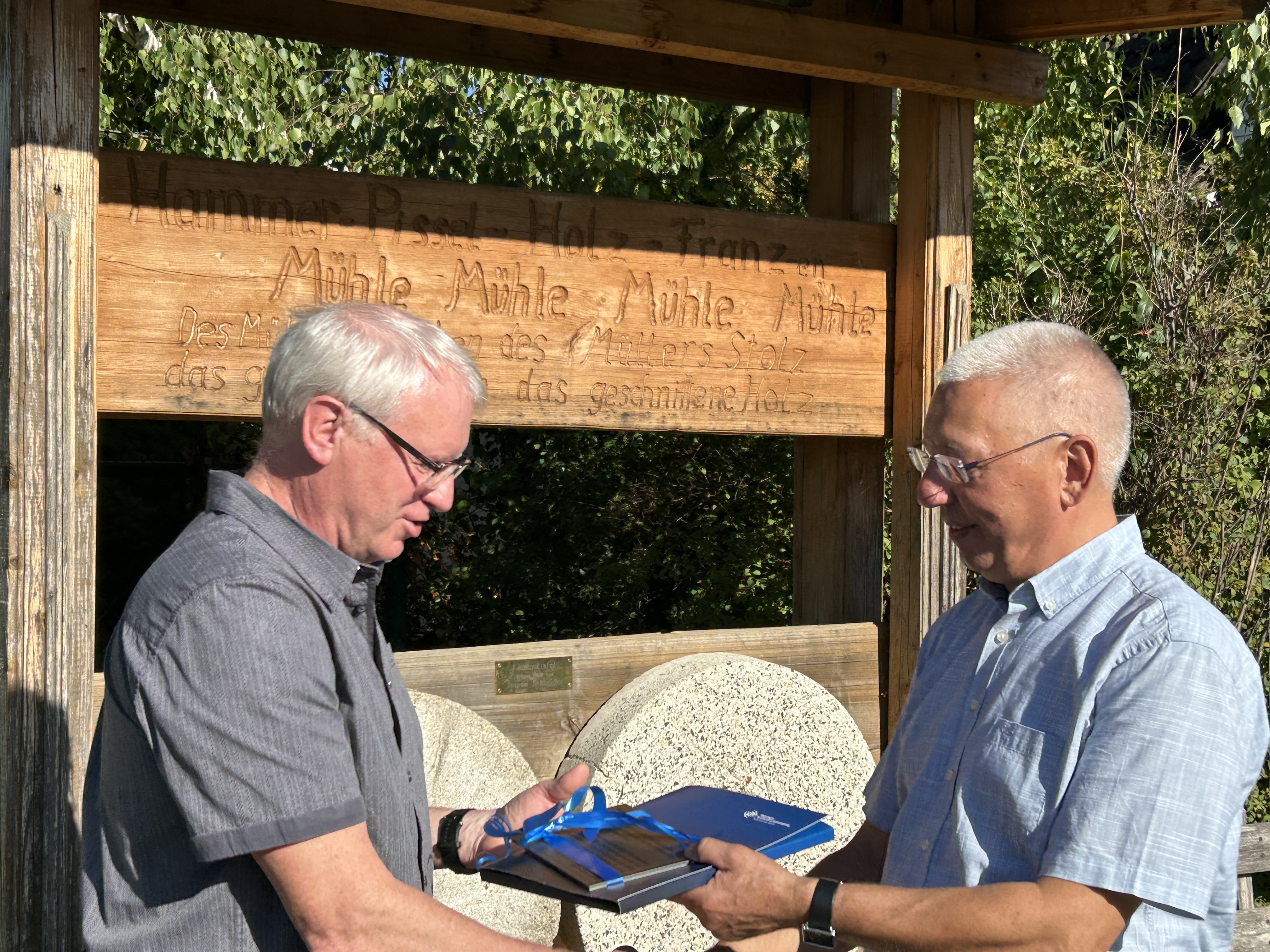 Mike Fritzsche ist links im Bild und erhält eine Plakette für die Mühlsteine von Quent Mehlhorn (rechts im Bild) übergeben. Beide stehen vor dem Mühlsteindenkmal.