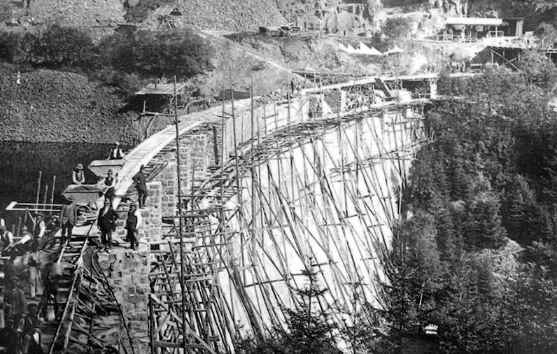 im Bau befindliche Staumauer mit Gerüst auf der Luftseite der Staumauer sowie die Arbeitenden sind auf der schwarz/weiß-Aufnahme