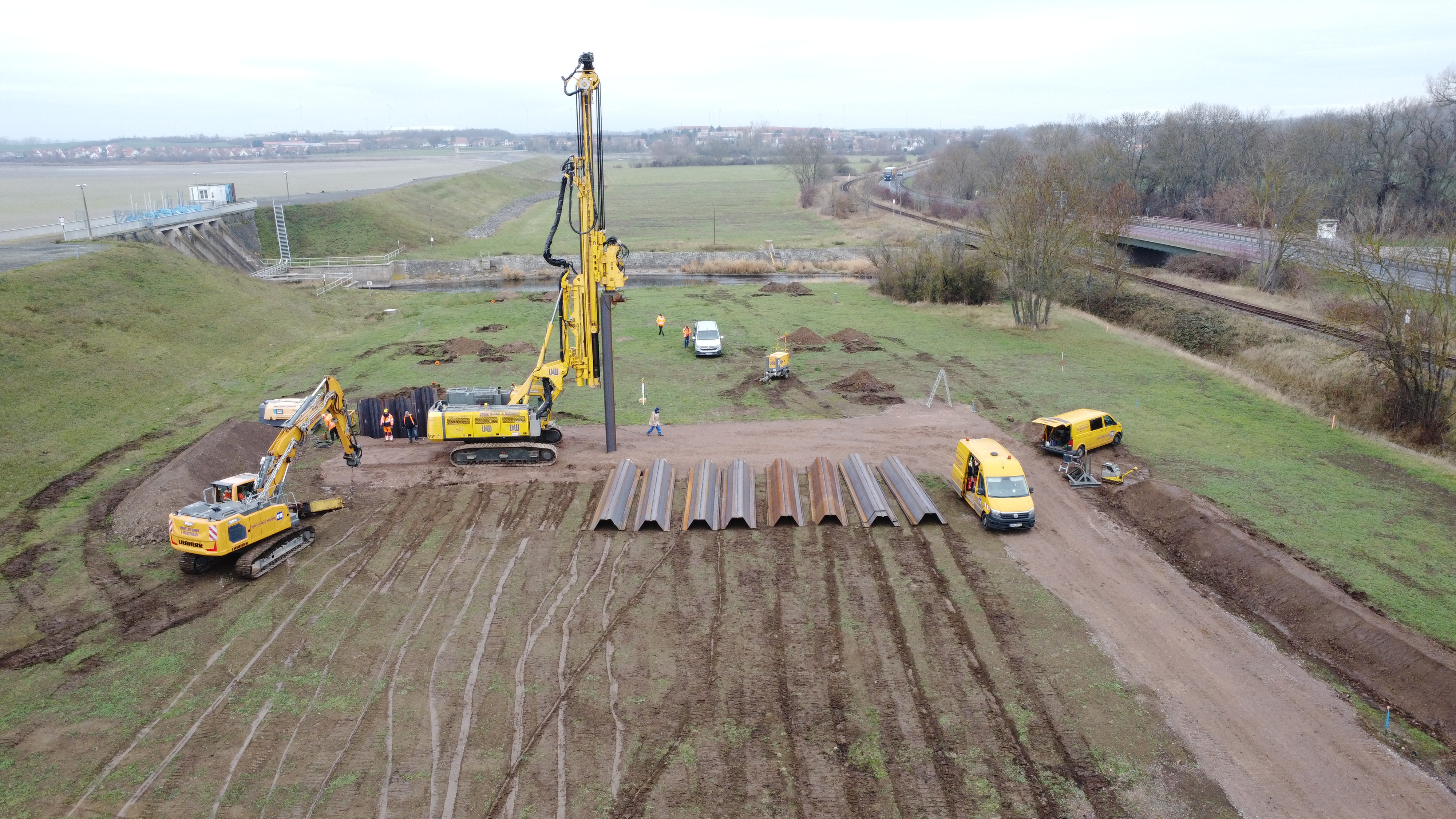 Eine Luftaufnahme des Probefelds am Hochwasserrückhaltebecken Straußfurt zeigt einen Teleskopmökler, auf der Erde liegende Spundwände und mehrere Baustellenfahrzeuge.