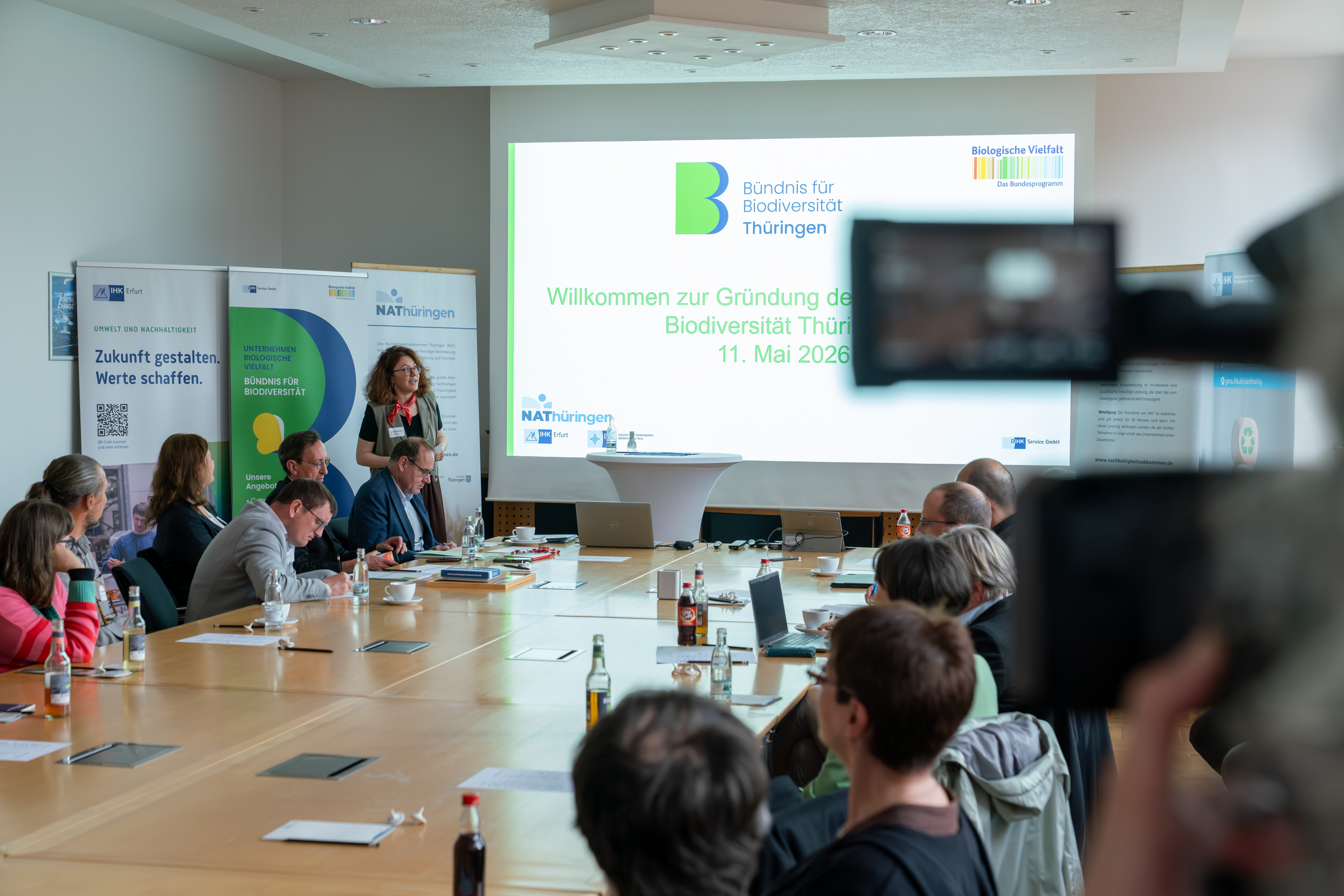 Eine Rednerin projiziert eine Folie mit der Überschrift "Gründungsveranstaltung Bündnis für Biodiversität Thüringen" an eine Wand, während an einer großen Tafel Bündnismitglieder sitzen und ihr gespannt lauschen.