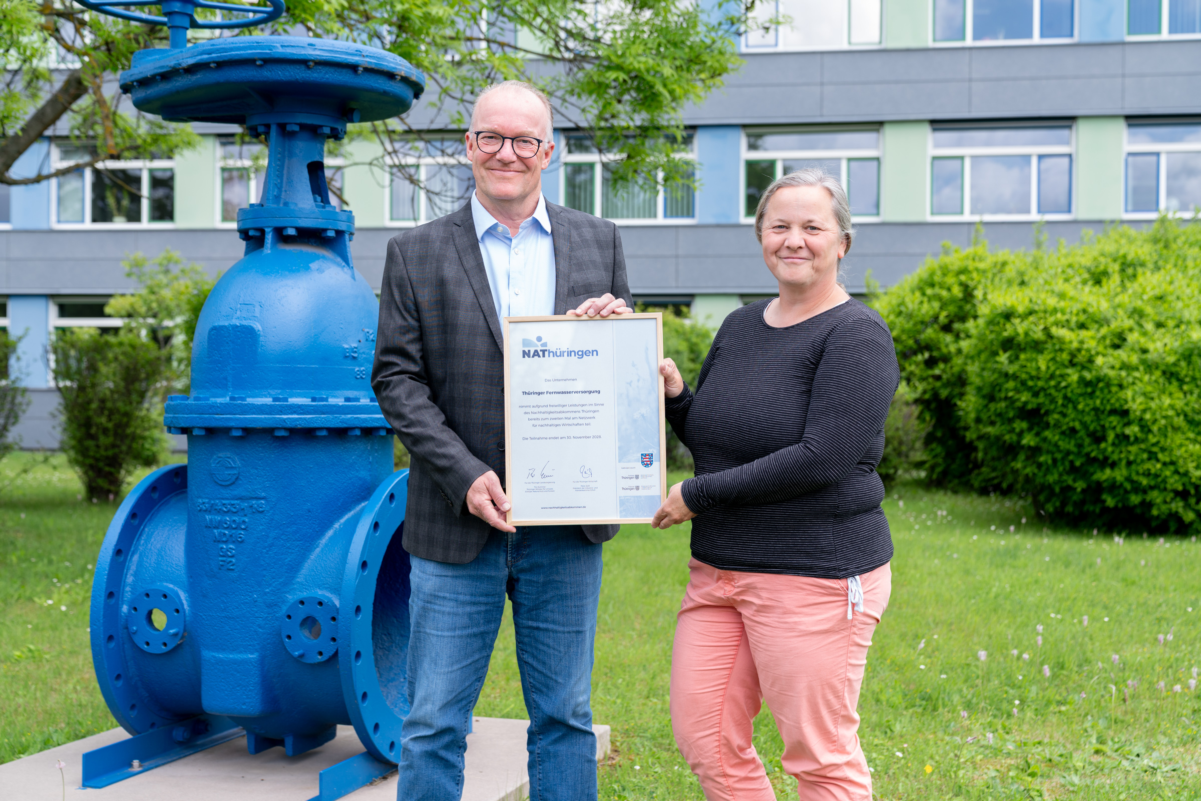 Thomas Dirkes, Geschäftsführer der Thüringer Fernwasserversorgung, nimmt vor dem Gebäude der Erfurter Hauptverwaltung eine Urkunde einer Mitarbeiterin von NAThüringen entgegen. Beide lächeln in die Kamera.