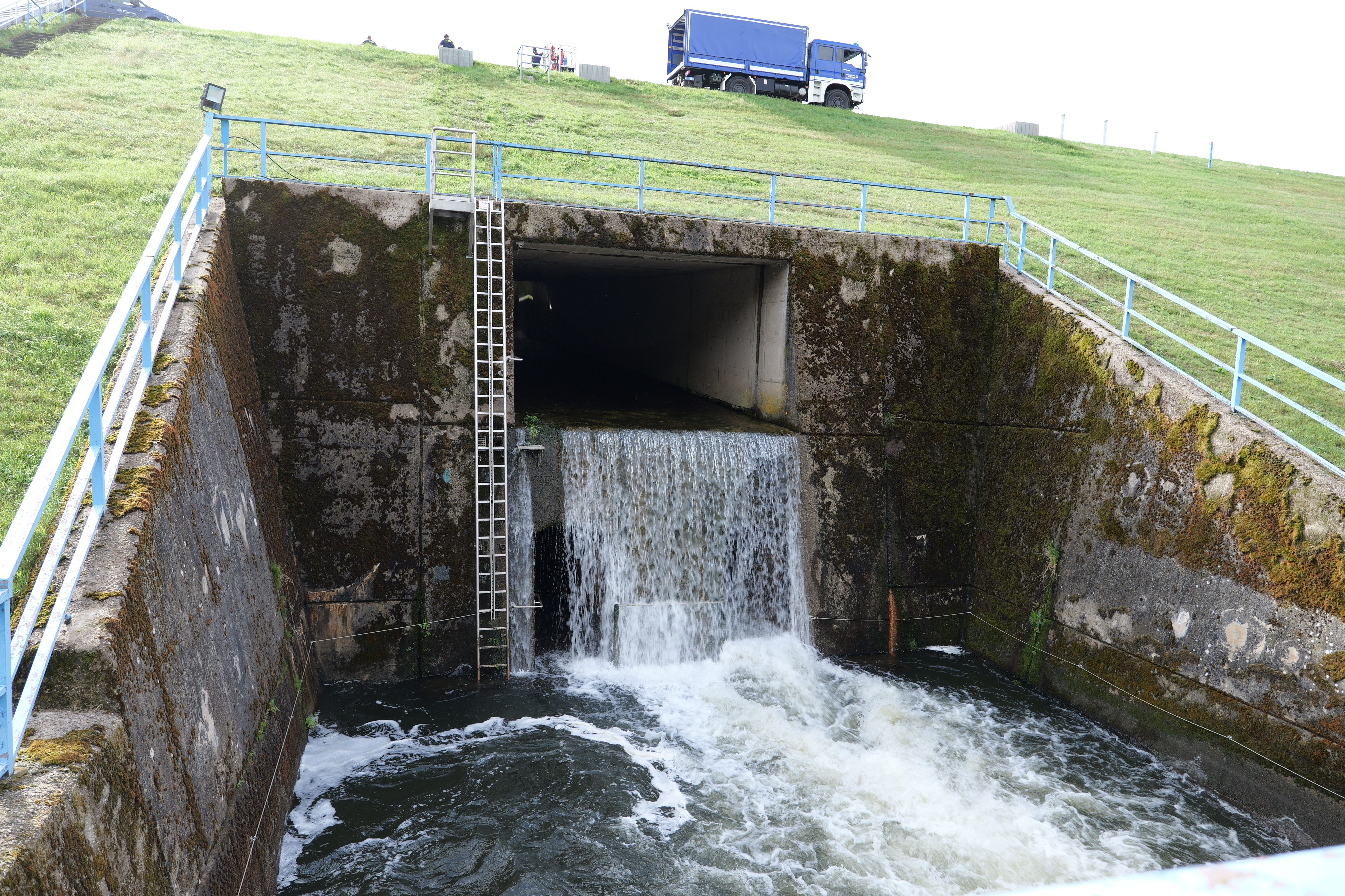 Wasser fließt aus der öffnung der Hochwasserentlastung in das Tosbecken. Auf der Dammkrone steht ein LKW des THW.